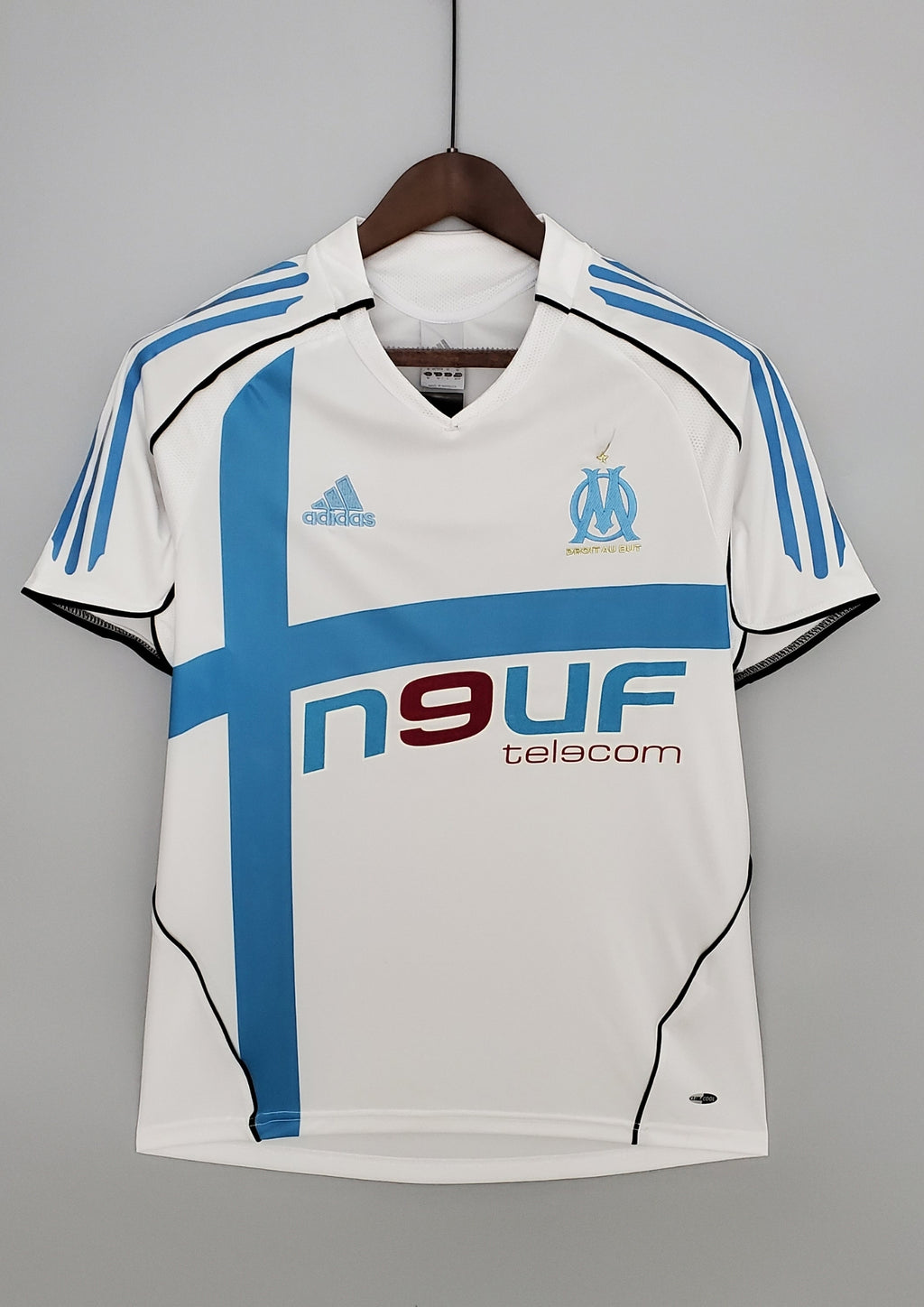 OM domicile 2006-2007 : maillot blanc Adidas avec une large croix bleu ciel asymétrique sur le torse, des liserés noirs incurvés et le sponsor Neuf Telecom. GoalKits