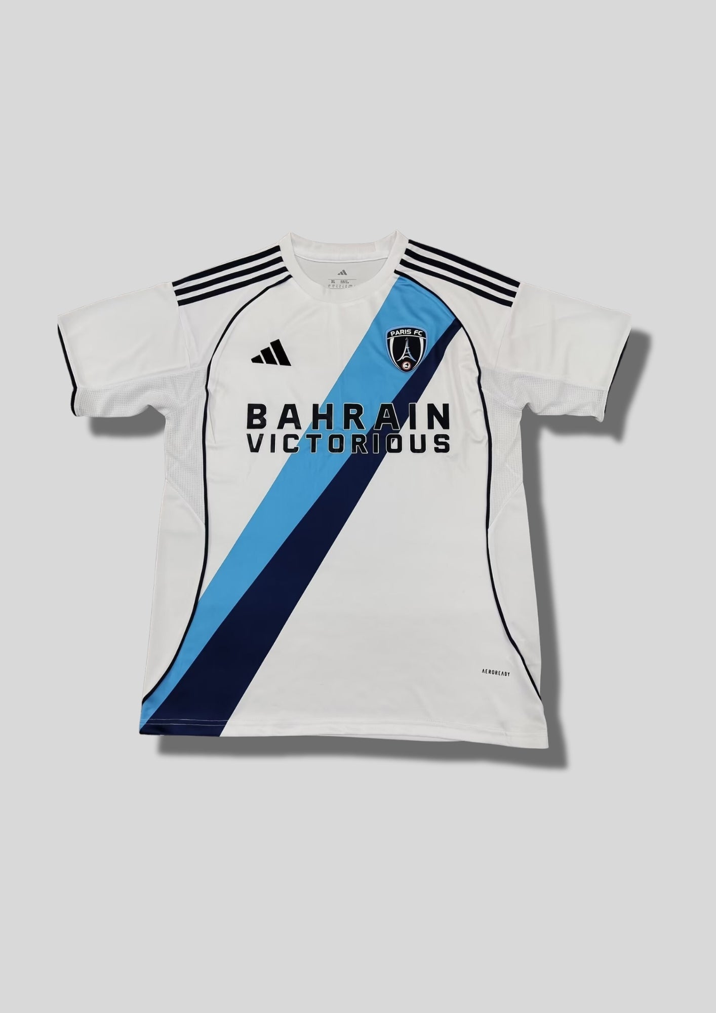 Paris FC extérieur 2024-2025 : maillot blanc Adidas avec une large bande diagonale bicolore (bleu ciel et bleu marine) passant sous le blason, sponsor Bahrain Victorious. GoalKits