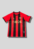 OGC Nice domicile 2024-2025 (120 ans) : maillot Le Coq Sportif aux rayures verticales rouges et noires traditionnelles, avec des logos et le sponsor INEOS dorés pour célébrer l'anniversaire du club. GoalKits