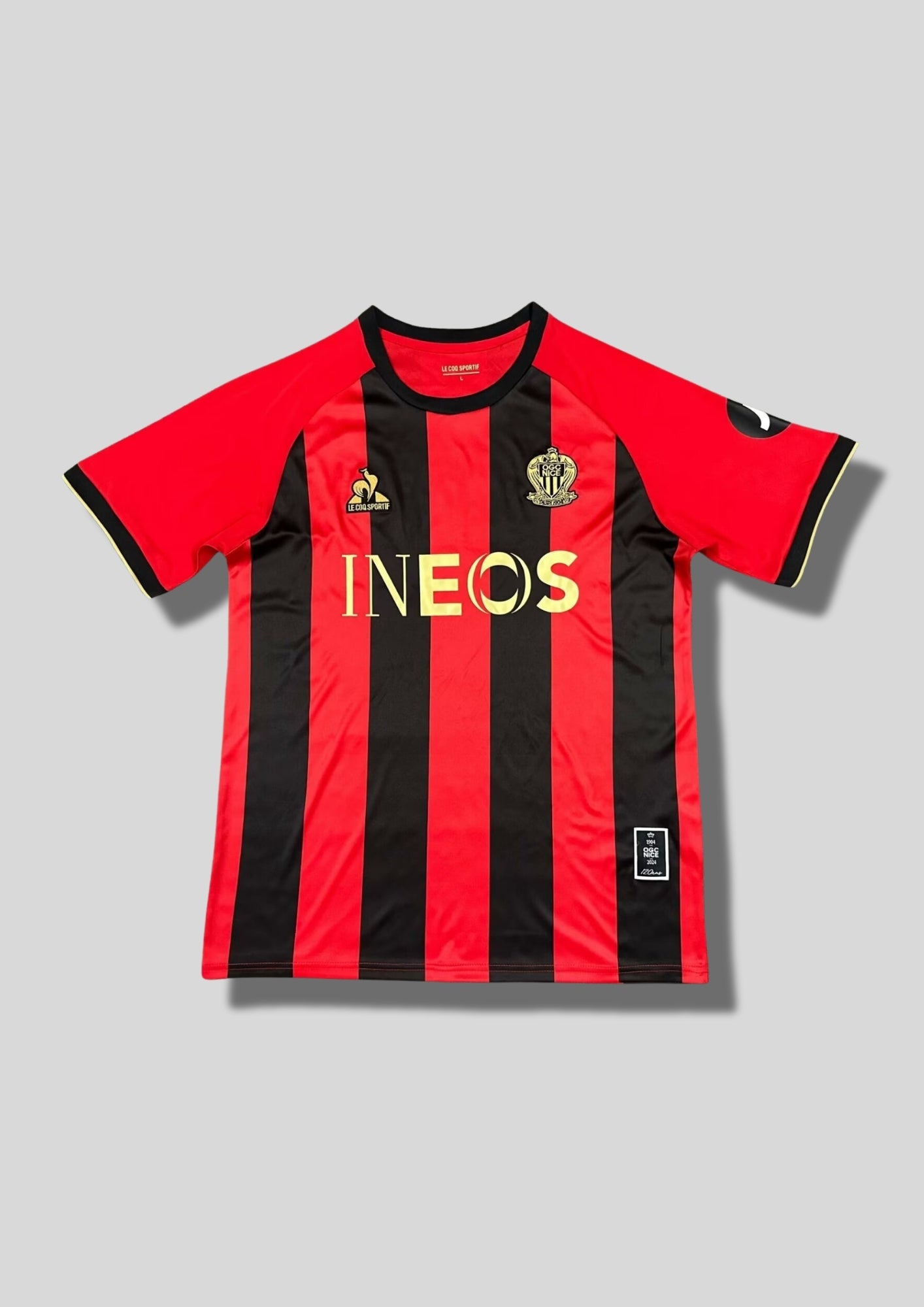 OGC Nice domicile 2024-2025 (120 ans) : maillot Le Coq Sportif aux rayures verticales rouges et noires traditionnelles, avec des logos et le sponsor INEOS dorés pour célébrer l'anniversaire du club. GoalKits