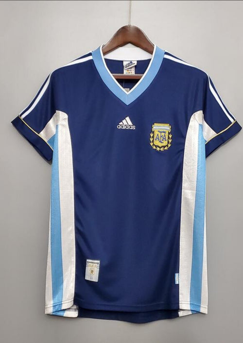 Argentine 1998 (Extérieur) Bleu marine, col en V ciel et blanc, bandes latérales tricolores, logo Adidas central blanc. GoalKits